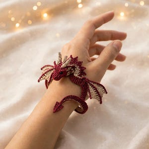 Puede incluir: Una pulsera de dragón tejida a ganchillo con un diseño en colores rojo oscuro y beige. La criatura tiene alas pequeñas, una cabeza detallada con cuernos y una cola larga y enroscada que rodea la muñeca. El diseño está hecho con puntadas finas.