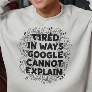 May include: A light gray sweatshirt with the text "Tired in Ways Google Cannot Explain" in a black, whimsical font. The design includes clocks, stars, and sleeping Z's. The sweatshirt has a crew neck and long sleeves.