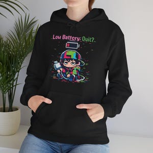 May include: Black hoodie featuring a colourful graphic of a cartoon character holding a plug, with the text "Low Battery: Quit?". The character is wearing a cap and is surrounded by pixelated lines. The hoodie has a front pocket and a hood.