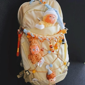 May include: A light beige backpack decorated with several small plush and plastic character charms attached to the front bungee cords and top pocket. The charms include a fuzzy yellow figure in a blue hat, a brown fuzzy creature, and a small boy figure.