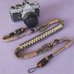 Puede incluir: Una cámara vintage plateada y negra con una lente negra se muestra con tres correas de muñeca para cámara sobre una superficie púrpura. Las correas son de color beige, verde oliva y negro, con hebillas negras ajustables y detalles naranjas.