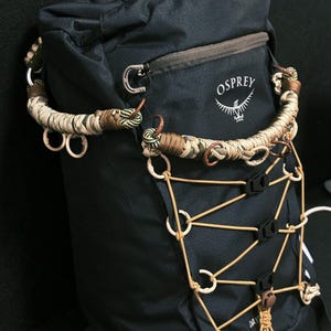 May include: A black Osprey backpack featuring a custom paracord handle wrap in tan, olive, and brown tones. The front includes a bungee cord lacing system with tan cords, small circular rings, and black plastic tension clips for securing gear.