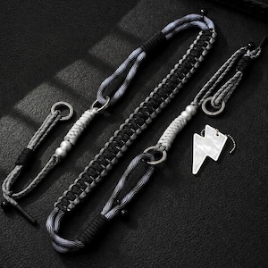 May include: A long, braided black and gray cord lanyard featuring a central woven section, silver-toned metal rings, and a white lightning bolt-shaped pendant. The accessory includes black cord accents, small silver beads, and a secure loop attachment.