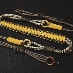 Op de afbeelding: Drie handgemaakte paracordarmbanden in geel, zwart, bruin en olijfgroen. Elke armband heeft een uniek geweven ontwerp met een cirkelvormige sluiting. De armbanden worden op een donkere ondergrond gepresenteerd.