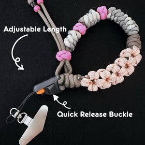 May include: A paracord lanyard featuring a braided pattern in shades of gray, white, and light pink, accented with bright pink knots. It includes a black quick release buckle with an orange button and a metal clip. Text reads Adjustable Length and Quick Release Buckle.