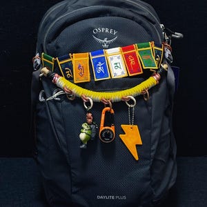 Può includere: Uno zaino nero Osprey Daylite Plus decorato con una fila di bandiere di preghiera tibetane colorate, un rivestimento in paracord giallo, una piccola statuetta di rana, una bussola a moschettone arancione e un ciondolo a fulmine giallo.