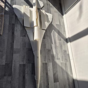 Può includere: Un modello bianco di un aereo supersonico Concorde. Il modello è appoggiato su un pavimento di legno grigio, con le ali e la coda ben visibili. La luce del sole proietta ombre sull'aereo e sul pavimento.