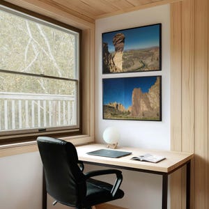 May include: A home office setup featuring a black leather office chair, a light wood desk with a dark laptop and an open book, a spherical white lamp, and two framed landscape photographs of rock formations hanging on a white wall.