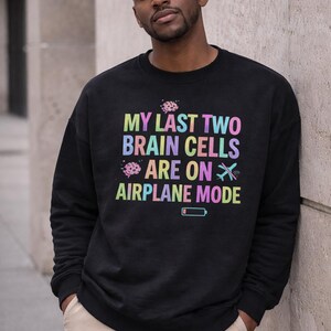 May include: Black sweatshirt with the text "MY LAST TWO BRAIN CELLS ARE ON AIRPLANE MODE" in rainbow colors. The design includes brain and airplane graphics, and a low battery icon. The person is wearing khaki pants.