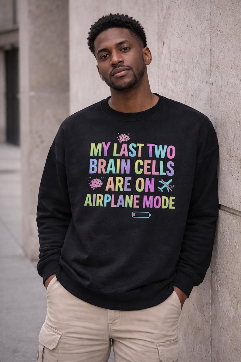 May include: Black sweatshirt with the text "MY LAST TWO BRAIN CELLS ARE ON AIRPLANE MODE" in rainbow colors. The design includes brain and airplane graphics, and a low battery icon. The person is wearing khaki pants.