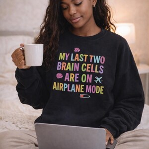 May include: Black sweatshirt with the text "MY LAST TWO BRAIN CELLS ARE ON AIRPLANE MODE" in rainbow colors. The design includes brain and airplane graphics, and a low battery icon. A woman is holding a white mug and using a laptop.