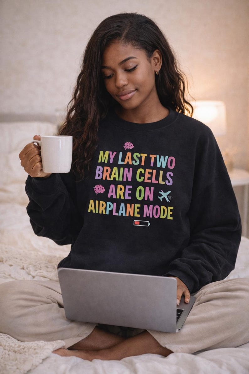 May include: Black sweatshirt with the text "MY LAST TWO BRAIN CELLS ARE ON AIRPLANE MODE" in rainbow colors. The design includes brain and airplane graphics, and a low battery icon. A woman is holding a white mug and using a laptop.