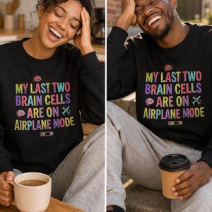 May include: Black long-sleeve shirt with the text "MY LAST TWO BRAIN CELLS ARE ON AIRPLANE MODE" in colorful letters. The shirt is worn by a person. A coffee mug is in the person's hand.