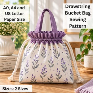 Può includere: Una borsa a secchiello con coulisse in tessuto color crema con un motivo ripetuto di rametti di lavanda viola e piccoli punti. La borsa presenta una parte superiore viola tinta unita con bordo arricciato, due manici viola e spesse corde bianche.