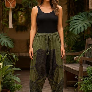 May include: A woman wearing a black tank top and olive green harem pants with a dark patch design. The pants have a drawstring waist. The background includes plants and a wooden bench, suggesting a relaxed, bohemian style.