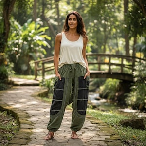 May include: A woman wearing a cream-colored tank top and olive green harem pants with black checkered pockets. The pants have a drawstring waist and are paired with brown sandals. The background features a stone path, a wooden bridge, and lush greenery.