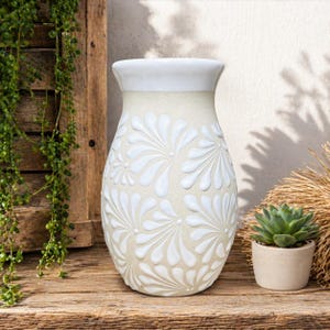May include: A cream-colored ceramic vase with a white floral pattern. The vase has a wide base, a narrow neck, and a white rim. The vase is displayed on a wooden surface with a small potted succulent.