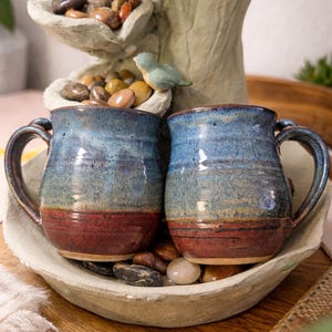 May include: Two handmade ceramic mugs with blue and red glaze. The mugs are displayed in a stone bowl with decorative pebbles. The mugs have a rustic, artisanal appearance, perfect for coffee or tea.