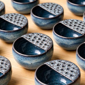 May include: A collection of blue ceramic bowls with a unique design. Each bowl has a dark blue interior and a textured, patterned section on one side. The bowls are arranged on a light-colored wooden surface, creating a visually appealing display.