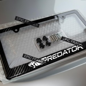 May include: A black license plate frame with a carbon fiber design and the word "PREDATOR" in white. The frame includes mounting hardware and the text "EPIC FRAMES".