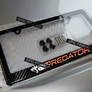 May include: Black license plate frame with a carbon fiber design and the word "PREDATOR" in orange. Includes mounting hardware. The frame is packaged in bubble wrap.