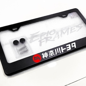 May include: Black license plate frame with white text that reads "EPIC FRAMES" and Japanese characters. Includes mounting hardware. The frame has a red and white logo and the text "神奈川トヨタ".