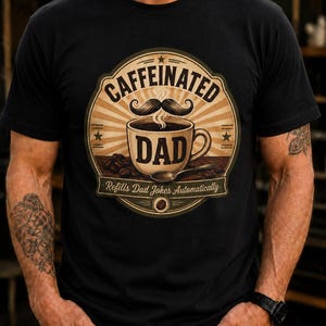 Op de afbeelding: Een zwart t-shirt met een cirkelvormige afbeelding van een koffiekop, een snor en de tekst "CAFFEINATED DAD" en "Refills Dad Jokes Automatically". Het ontwerp bevat een zonnestraal-achtergrond, koffiebonen en een lepel.