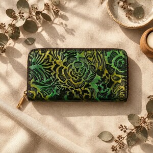 May include: A rectangular wallet featuring a vibrant green and black embossed floral pattern across its surface. The wallet is secured with a dark brown zip that runs along the top and sides, finished with a polished gold-tone metal zip pull.