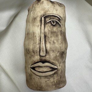 Può includere: Un vaso in ceramica beige con un design a volto scolpito. Il vaso ha una forma rettangolare con una superficie strutturata e presenta un singolo occhio, un naso lungo e un paio di labbra. Il vaso è un oggetto decorativo.