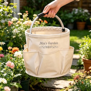 May include: A beige canvas garden tote with a silver handle and black grip is held up in front of a garden. The tote has multiple pockets and the text "Mia's Garden" printed on the front.