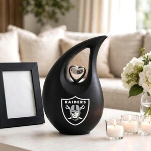 Op de afbeelding: Een matzwarte, traanvormige urn met een zilverkleurig hartvormig accent bij de opening aan de bovenzijde en het logo van het Raiders football-team in wit, zwart en zilver op de voorzijde van het object.