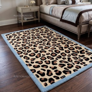 May include: A rectangular area rug featuring a repeating leopard print pattern in shades of tan, brown, and black. The rug is finished with a solid light blue border that frames the animal print design. The text RUSTICRUGCOO is visible on the rug.