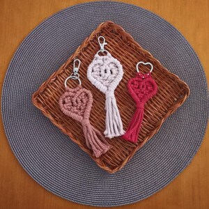 Puede incluir: Tres llaveros de macramé en forma de corazón en tonos rosa, blanco y rojo, cada uno con un cierre plateado y una borla. Los llaveros se exhiben en una cesta tejida sobre un salvamanteles gris, con una mesa de madera al fondo.