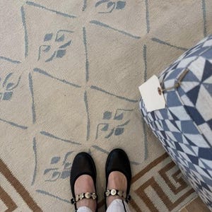 May include: A pair of black leather Mary Jane shoes featuring a strap adorned with metallic floral embellishments. The shoes are worn with white trousers, positioned on a beige rug with a blue diamond pattern and a blue and white geometric ottoman.