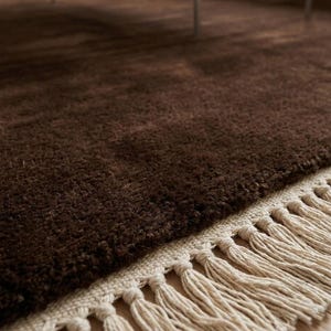 Premium chocolate brown hand-knotted rug made of 100% New Zealand wool, featuring a solid contemporary design with contrasting fringed edges, styled under a mid-century coffee table in a modern minimalist living room.