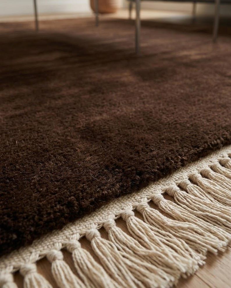 Premium chocolate brown hand-knotted rug made of 100% New Zealand wool, featuring a solid contemporary design with contrasting fringed edges, styled under a mid-century coffee table in a modern minimalist living room.