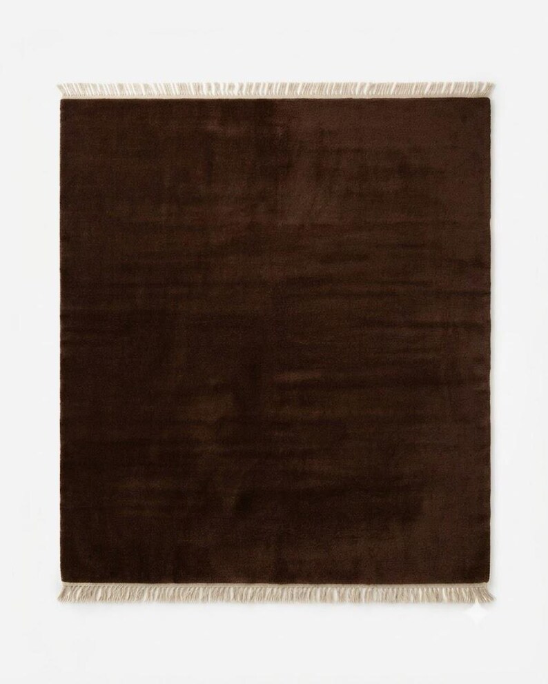 Premium chocolate brown hand-knotted rug made of 100% New Zealand wool, featuring a solid contemporary design with contrasting fringed edges, styled under a mid-century coffee table in a modern minimalist living room.