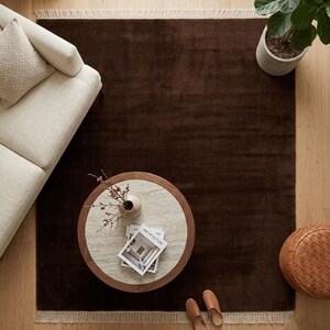 Premium chocolate brown hand-knotted rug made of 100% New Zealand wool, featuring a solid contemporary design with contrasting fringed edges, styled under a mid-century coffee table in a modern minimalist living room.