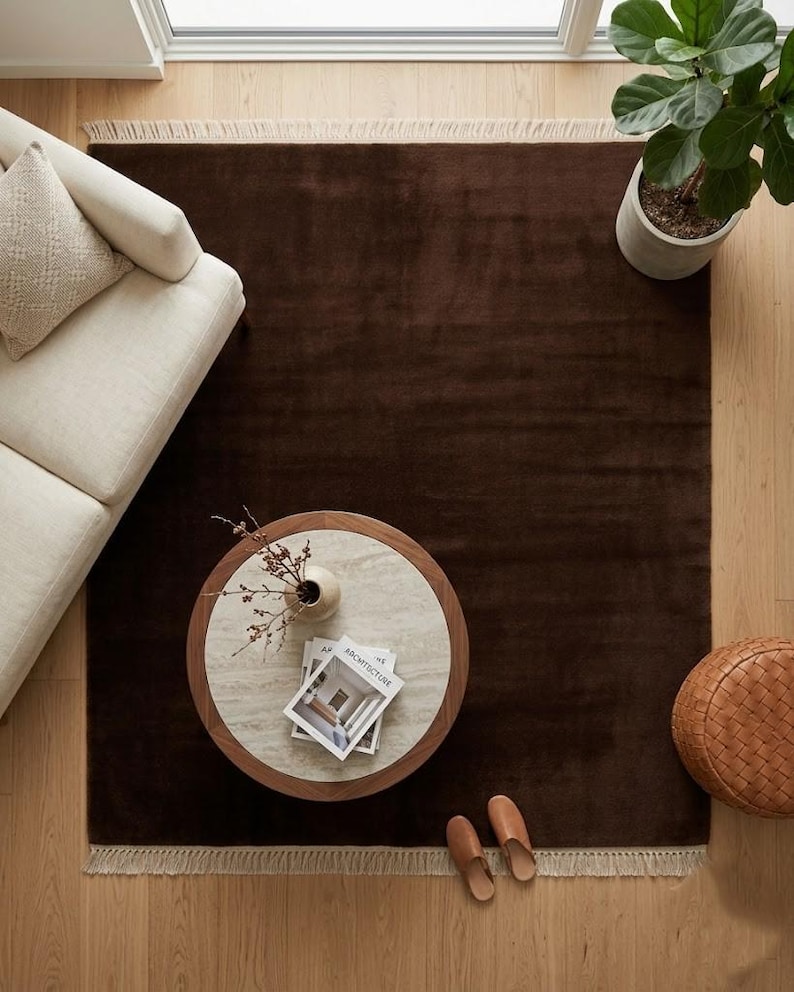 Premium chocolate brown hand-knotted rug made of 100% New Zealand wool, featuring a solid contemporary design with contrasting fringed edges, styled under a mid-century coffee table in a modern minimalist living room.