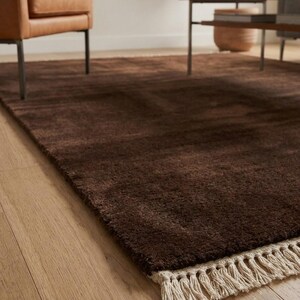 Premium chocolate brown hand-knotted rug made of 100% New Zealand wool, featuring a solid contemporary design with contrasting fringed edges, styled under a mid-century coffee table in a modern minimalist living room.