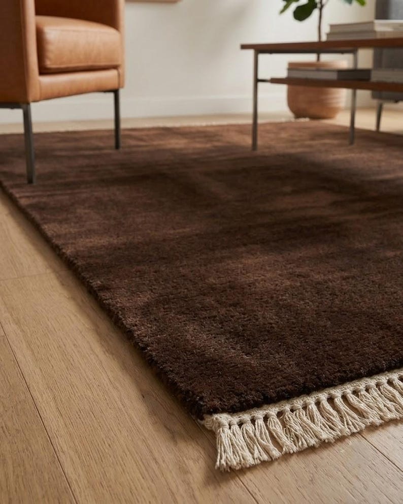 Premium chocolate brown hand-knotted rug made of 100% New Zealand wool, featuring a solid contemporary design with contrasting fringed edges, styled under a mid-century coffee table in a modern minimalist living room.