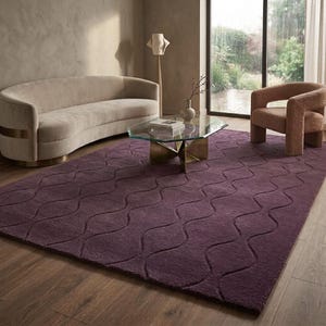 Könnte beinhalten: Ein großer, rechteckiger, tiefvioletter Teppich mit einem wellenförmigen Muster dominiert einen Wohnraum. Der Teppich liegt auf einem Holzboden, mit einem geschwungenen beigen Sofa und einem modernen Sessel. Ein Glastisch steht im Zentrum.