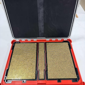 May include: Open red case with a clear lid, revealing a black interior and multiple compartments filled with small, gold-colored cylindrical objects. The case is designed for organization and storage.