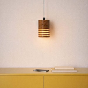 Pu&ograve; includere: Un lampadario cilindrico con finitura in legno. La luce ha fessure orizzontali che emettono un bagliore caldo. L'apparecchio &egrave; appeso a un cavo nero. Un mobile giallo, uno smartphone e dei libri sono in primo piano.