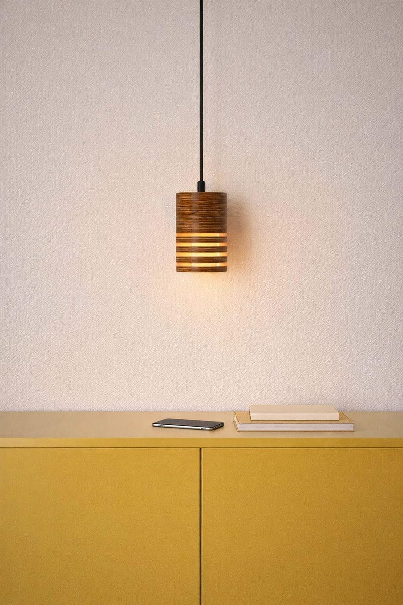 Pu&ograve; includere: Un lampadario cilindrico con finitura in legno. La luce ha fessure orizzontali che emettono un bagliore caldo. L'apparecchio &egrave; appeso a un cavo nero. Un mobile giallo, uno smartphone e dei libri sono in primo piano.