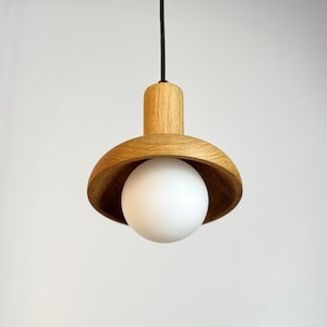 Pode incluir: Uma moderna luminária pendente com uma sombra de madeira e uma lâmpada de globo branca. A luz pende de um cabo preto. A sombra de madeira tem um acabamento natural e a lâmpada é redonda.