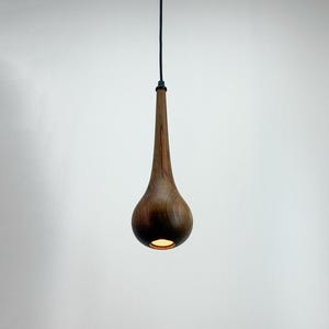 May include: A teardrop-shaped wooden pendant light fixture. The light fixture is suspended from a dark green cord. The wood has a rich, dark brown color, and the light shines from the bottom.