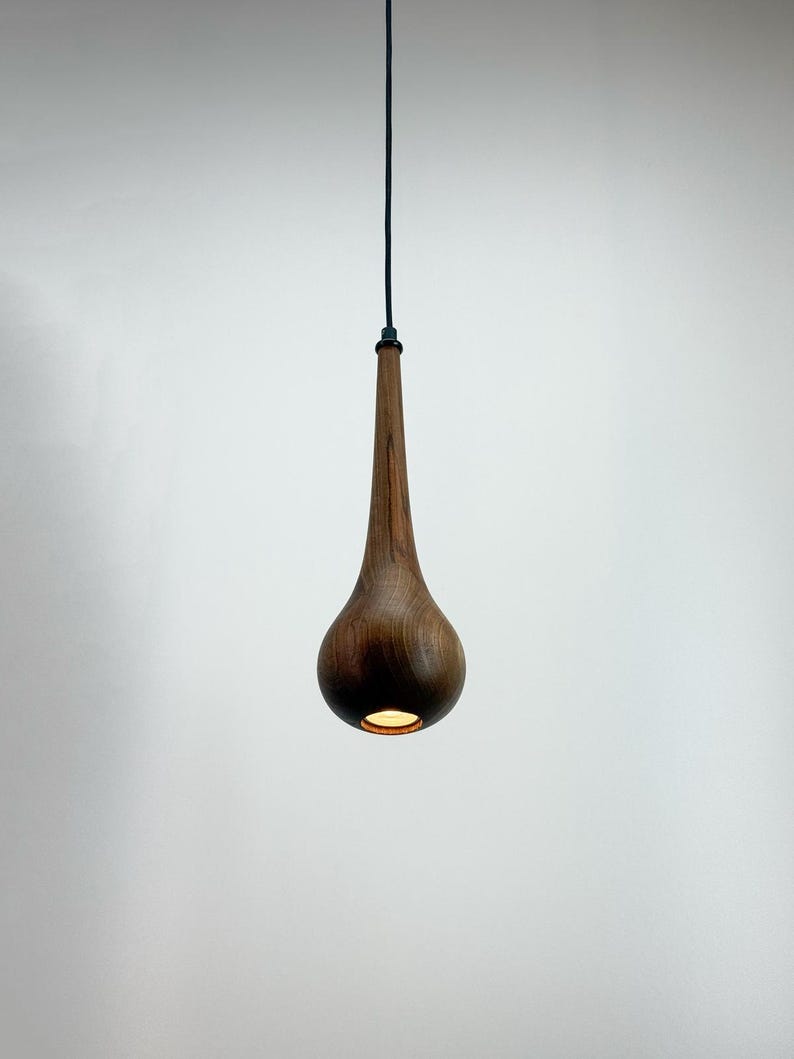 May include: A teardrop-shaped wooden pendant light fixture. The light fixture is suspended from a dark green cord. The wood has a rich, dark brown color, and the light shines from the bottom.