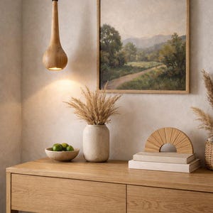 May include: A wooden pendant light hangs above a wooden console table. The table holds a vase with dried plants, a bowl of limes, a stack of books, and a decorative wooden semi-circle. A framed landscape painting hangs on the wall.