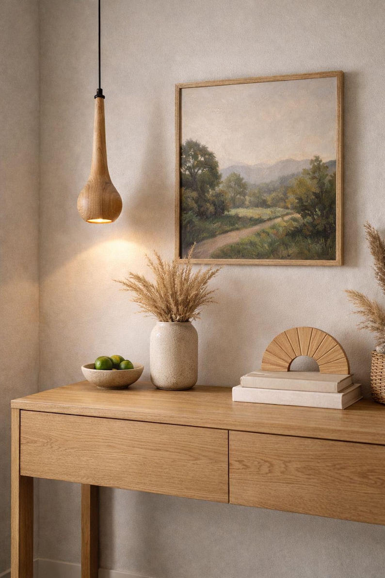 May include: A wooden pendant light hangs above a wooden console table. The table holds a vase with dried plants, a bowl of limes, a stack of books, and a decorative wooden semi-circle. A framed landscape painting hangs on the wall.